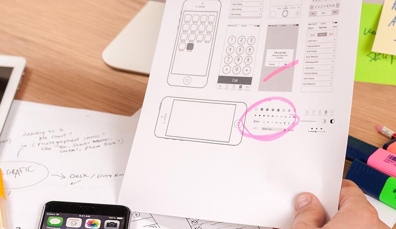 Photo montrant le design d'interface pour mobile, une main dans le coin droit et bas de la photo, tient une feuille de papier blanche avec des protype de pages d'une application pour smartphone